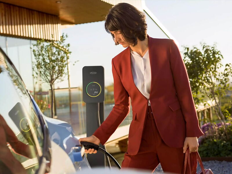 Frau in rotem Businessanzug, die vor dem Haus den Stromstecker ans E-Auto anschliesst