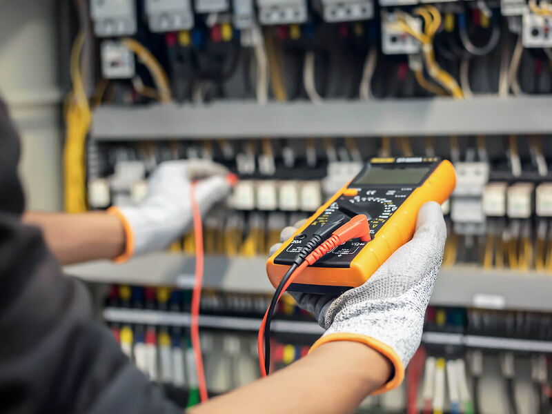 Hände mit Handschuhen, die an einem Stromtableau mit orangem Gerät und Kabeln den Strom messen