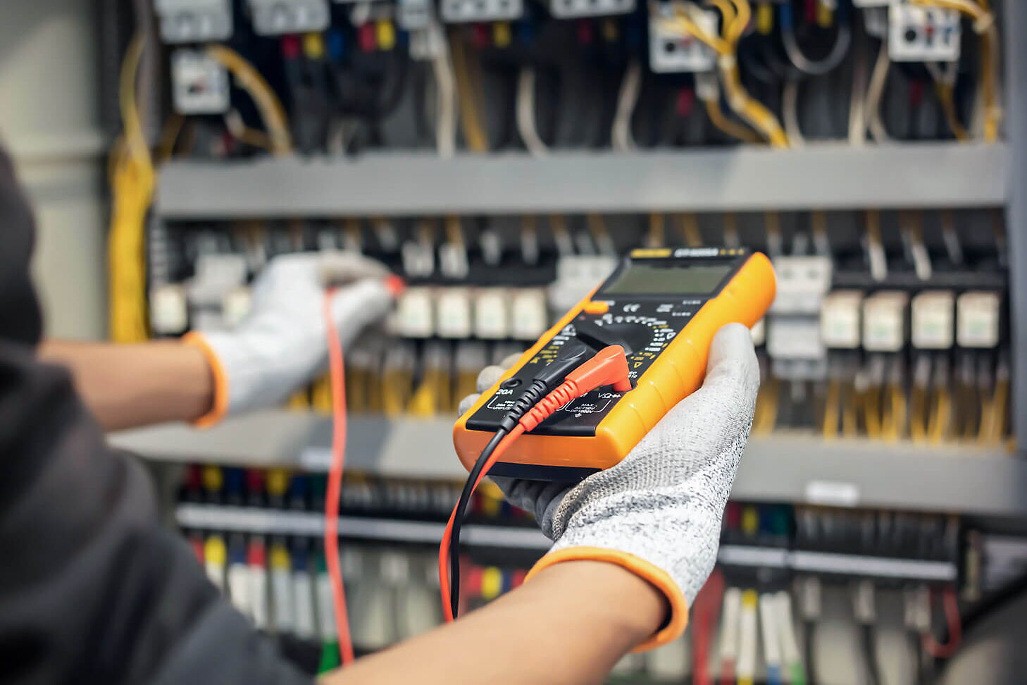 Hände mit Handschuhen, die an einem Stromtableau mit orangem Gerät und Kabeln den Strom messen